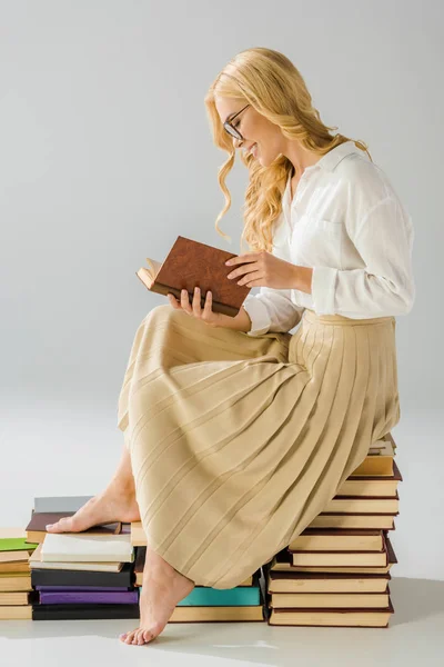 Atractiva mujer descalza sonriente en gafas leyendo y sentada en libros - foto de stock