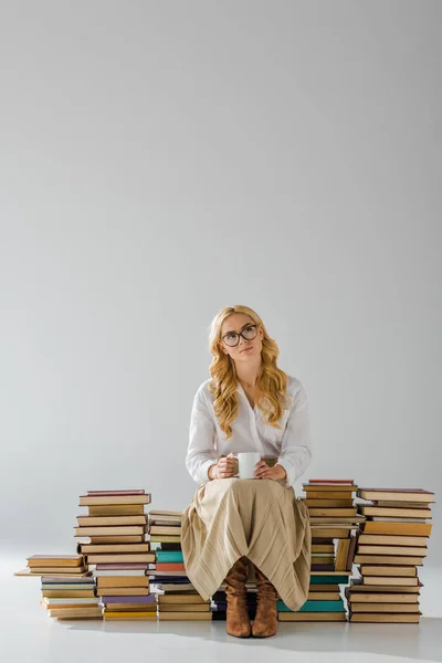 Donna attraente in occhiali con tazza seduta su mucchio di libri retrò — Foto stock