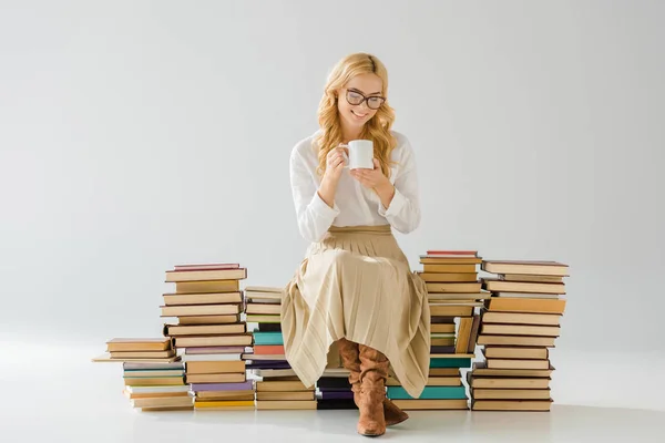 Attractive woman in glasses drinking coffee and sitting on pile of retro books — Stock Photo