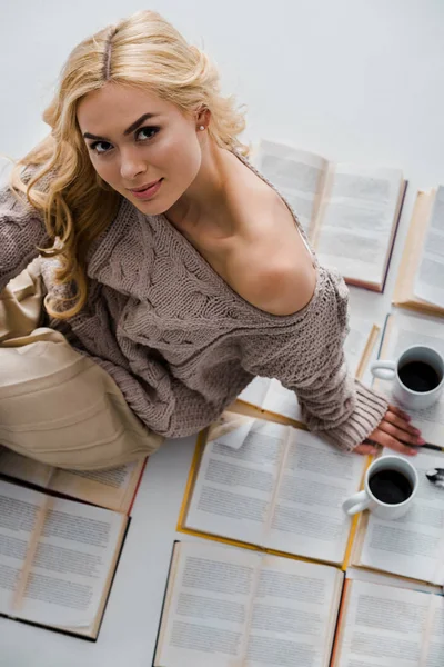 Primer plano de la mujer sentada en libros abiertos con tazas de café aisladas en gris - foto de stock