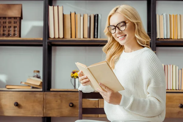 Primo piano di bella donna in occhiali seduta e lettura libro in soggiorno — Foto stock