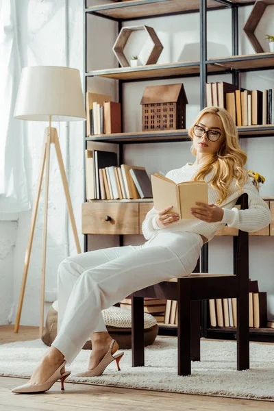 Hermosa mujer adulta en gafas sentado en la silla y libro de lectura - foto de stock