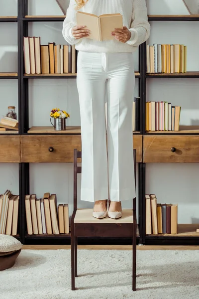 Close up of adult woman standing on chair in living room and reading book — Stock Photo