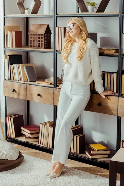 Attraente donna in occhiali in piedi vicino rack con libri in soggiorno — Foto stock