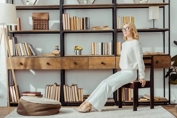 Adult blonde woman in glasses sitting near rack with books — Stock Photo