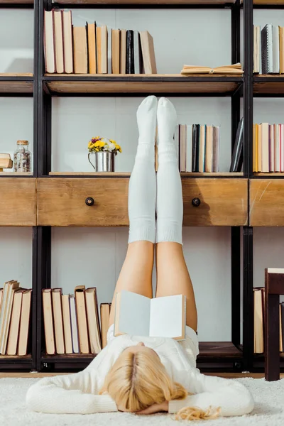 Woman laying on carpet and reading book with legs on rack — Stock Photo