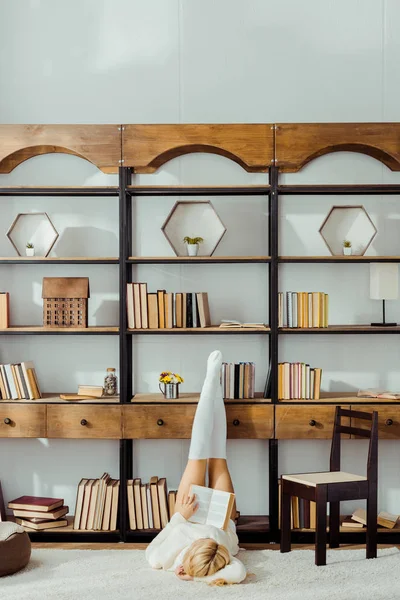 Femme posée sur le tapis et le livre de lecture avec les jambes en chaussettes blanches sur rack — Photo de stock