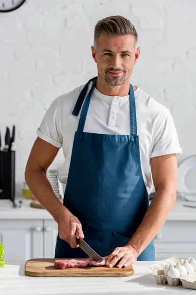 Mature homme souriant coupant steak sur planche en bois — Photo de stock