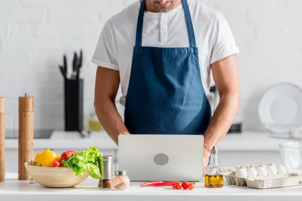 Visão cortada de homem em avental trabalhando no laptop na cozinha — Fotografia de Stock