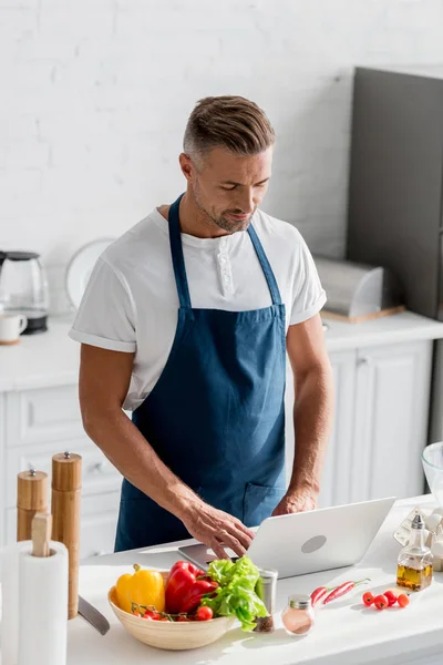 Homem bonito em avental de pé na cozinha e trabalhando no laptop — Fotografia de Stock