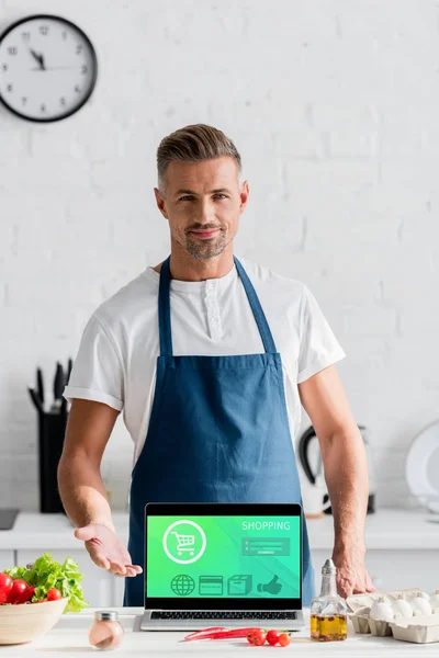 Homem adulto bonito de pé com ilustração de compras on-line na tela do laptop na mesa da cozinha — Fotografia de Stock