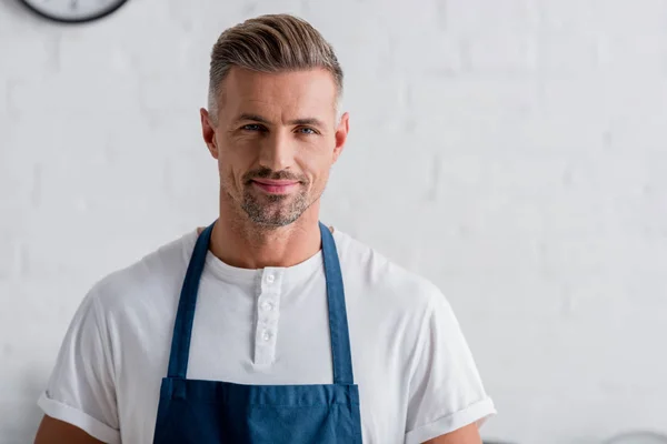 Adult smiling man in apron standing at kitchen — Stock Photo