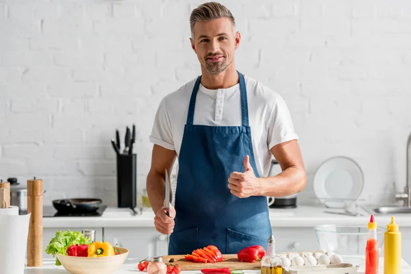 Hombre mostrando los pulgares hacia arriba con cuchillo en la mano de pie en la cocina — Stock Photo