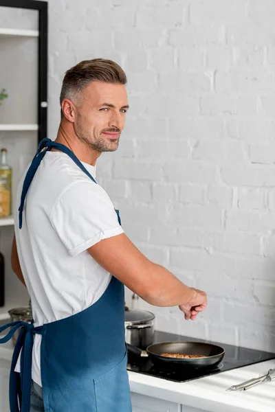 Handsome man in apron salting steak on pan — Stock Photo