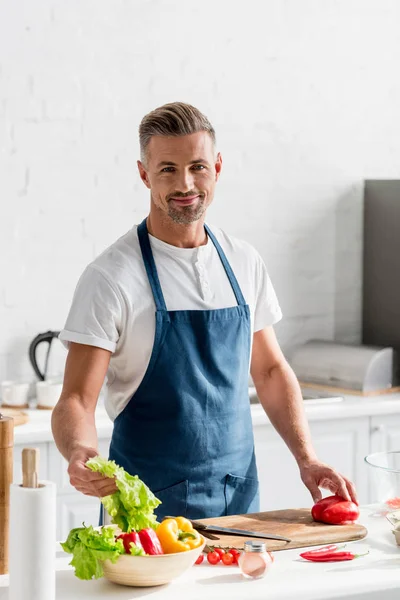 Uomo adulto in piedi in cucina con ingredienti insalata sul tavolo — Foto stock