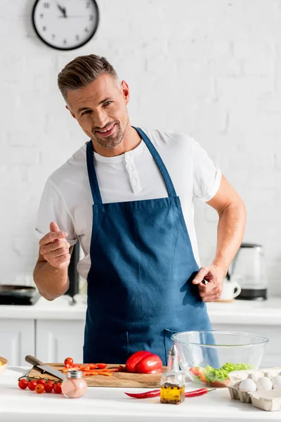 Bell'uomo sorridente in grembiule che cucina cibo in cucina — Foto stock