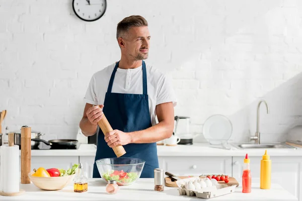 Schöner erwachsener Mann in Schürze salzt Salat in der Küche — Stockfoto