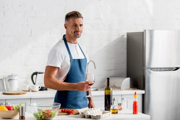 Hombre adulto en delantal con copa de vino en la cocina - foto de stock