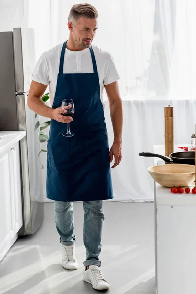 Hombre en delantal caminando a través de la cocina con vaso de vino - foto de stock