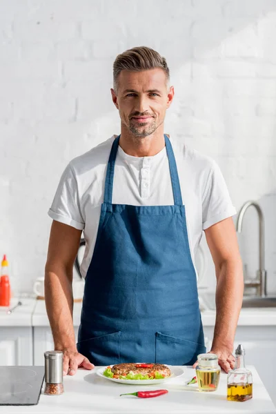 Handsome male cooker standing at kitchen with prepared dinner — Stock Photo