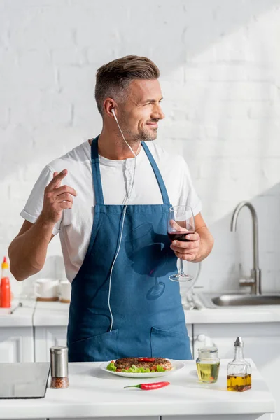 Adult man in apron with glass of wine and cooked steak listening to music — Stock Photo