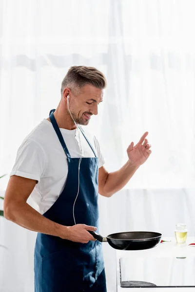 Улыбающийся мужчина с наушниками и сковородкой в руке, слушающий музыку на кухне — стоковое фото