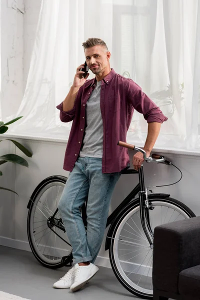 Мужчина разговаривает на смартфоне и опирается на велосипед в домашнем офисе — стоковое фото