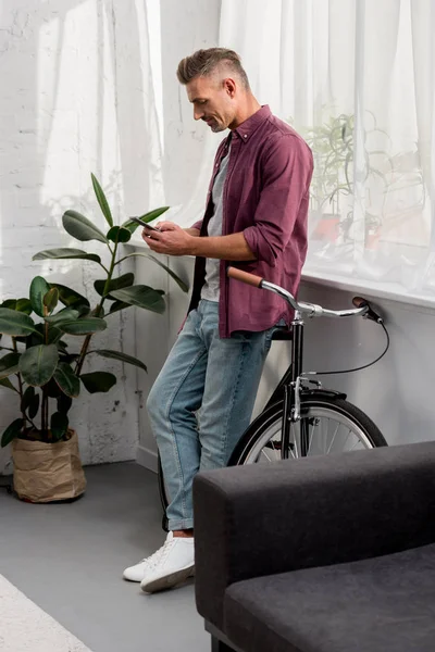 Homme utilisant un smartphone tout en s'appuyant sur le vélo au bureau à la maison — Photo de stock