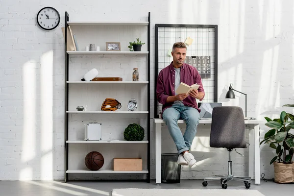 Hombre sentado en la mesa y leyendo libro en casa oficina - foto de stock