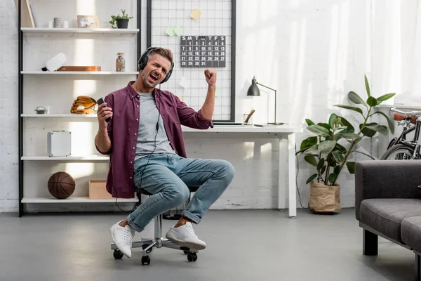Hombre feliz escuchando música y cantando mientras está sentado en la silla en la oficina en casa - foto de stock
