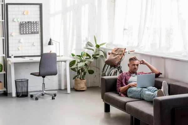 Nachdenklicher Mann, der auf dem Sofa liegt und zu Hause im Büro am Laptop arbeitet — Stockfoto