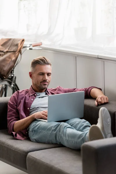 Freelancer bonito que coloca no sofá e trabalhando no laptop no escritório em casa — Fotografia de Stock
