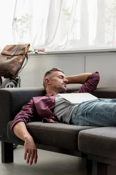 Человек лежит на диване с ноутбуком и спит в домашнем офисе — стоковое фото