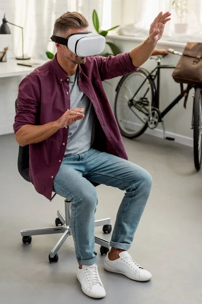 Человек сидит на стуле и жестикулирует в гарнитуре виртуальной реальности в домашнем офисе — стоковое фото