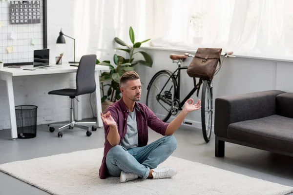 Концентрированный мужчина, сидящий на полу и практикующий йогу в домашнем офисе — стоковое фото