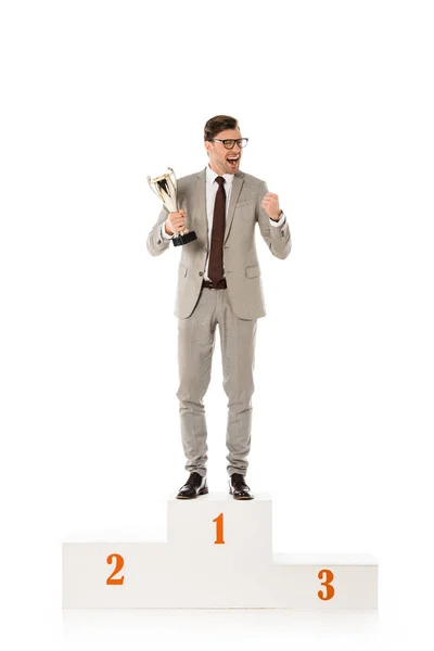 Handsome excited businessman with trophy cup standing on winners podium isolated on white — Stock Photo