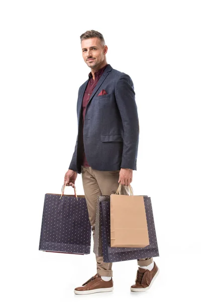 Cheerful adult stylish man in jacket holding colorful shopping bags isolated on white — Stock Photo