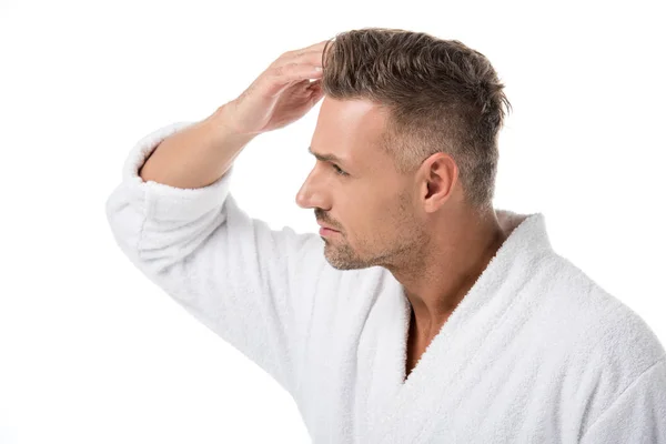 Vista lateral del hombre adulto molesto en albornoz comprobándose a sí mismo para la pérdida de cabello aislado en blanco - foto de stock