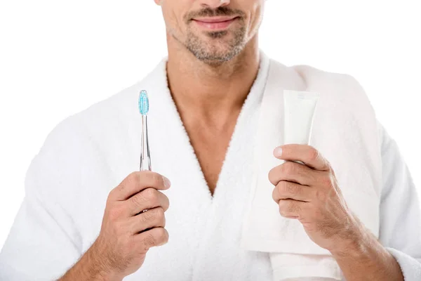 Vista parcial del hombre en albornoz con toalla sobre el hombro sosteniendo pasta de dientes y cepillo de dientes aislados en blanco - foto de stock