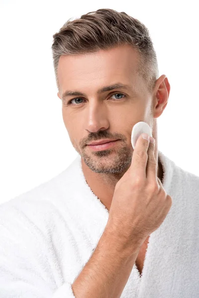 Retrato de hombre guapo en albornoz aplicando tónico por almohadilla de algodón en la cara aislada en blanco - foto de stock