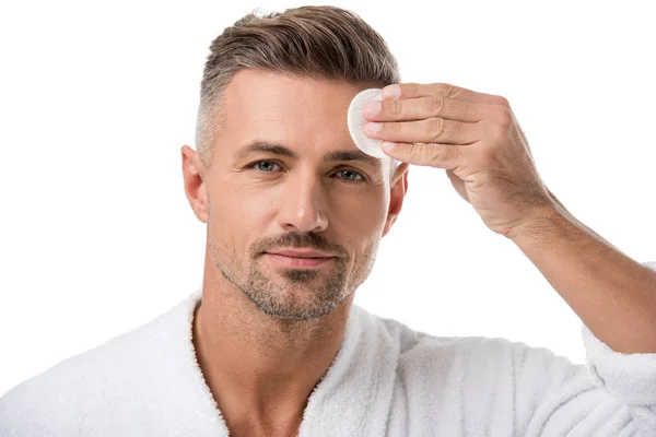 Retrato de hombre guapo en albornoz aplicando tónico por almohadilla de algodón en la cara aislada en blanco - foto de stock