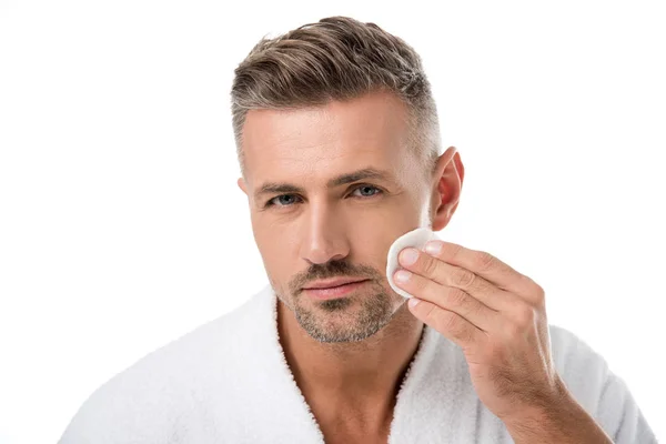 Portrait of handsome man in bathrobe applying tonic by cotton pad on face isolated on white — Stock Photo