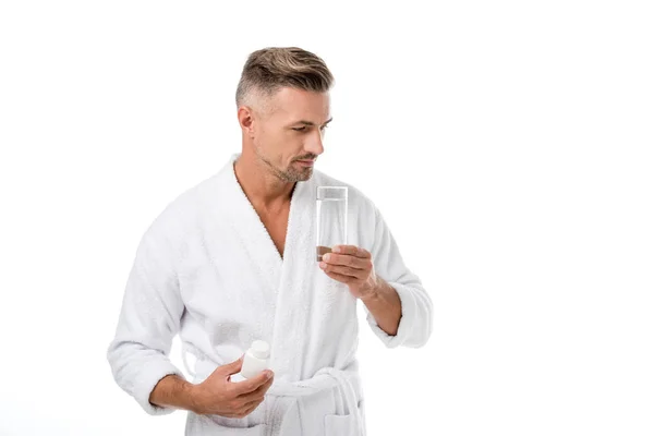Homme adulte joyeux en peignoir montrant des vitamines et tenant verre d'eau isolé sur blanc — Photo de stock