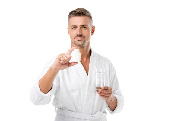 Hombre adulto alegre en albornoz mostrando vitaminas y sosteniendo vaso de agua aislado en blanco - foto de stock