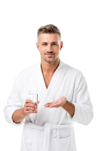 Hombre sonriente en albornoz sosteniendo píldoras y vaso de agua aislado en blanco - foto de stock