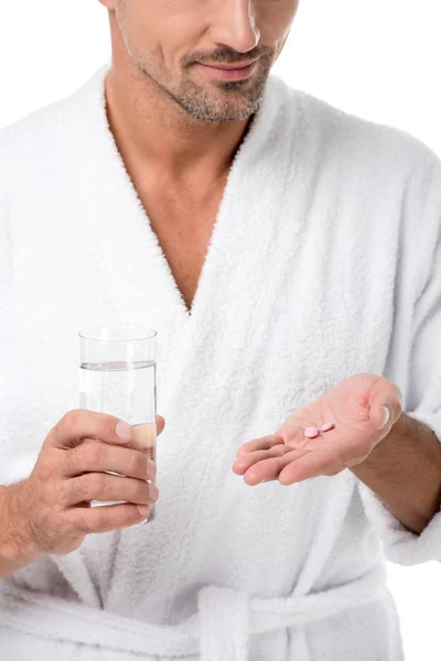 Partial view of adult man in bathrobe holding pills and glass of water isolated on white — Stock Photo