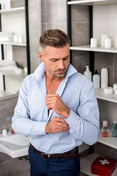 Confident adult businessman buttoning up blue shirt in bathroom at home — Stock Photo