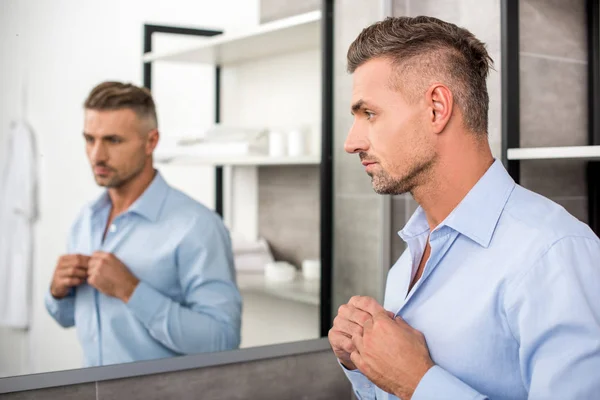 Selective focus of adult businessman buttoning up blue shirt in bathroom at home — Stock Photo