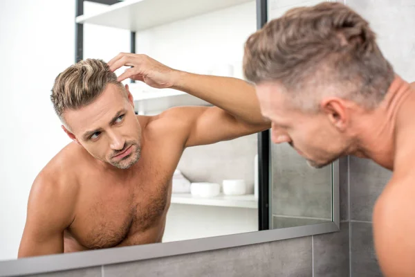 Красивый взрослый мужчина регулирует прическу, глядя на зеркало в ванной комнате — стоковое фото