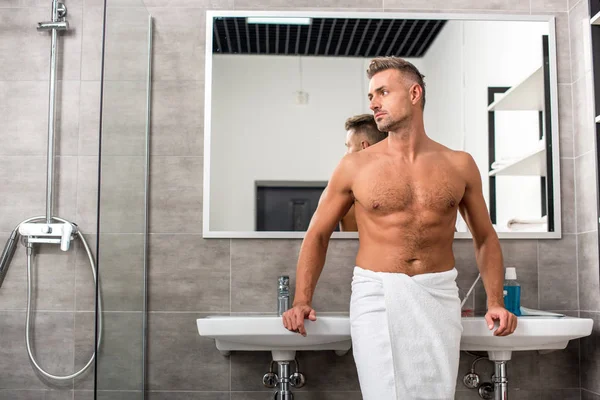 Confident muscular shirtless man in towel posing near sinks in bathroom — Stock Photo